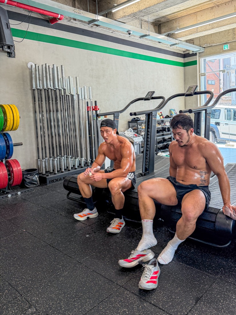 Barbells and bumper plates stored along the wall, two people resting
